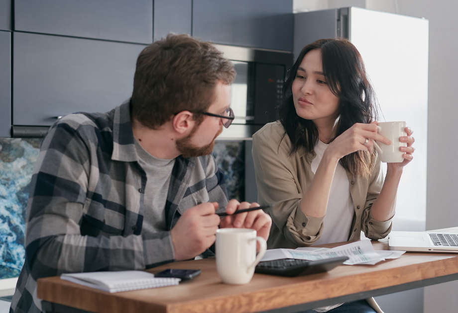 Man and woman sitting next to each other at a table in a kitchen, drinking coffee and engaged in conversation
